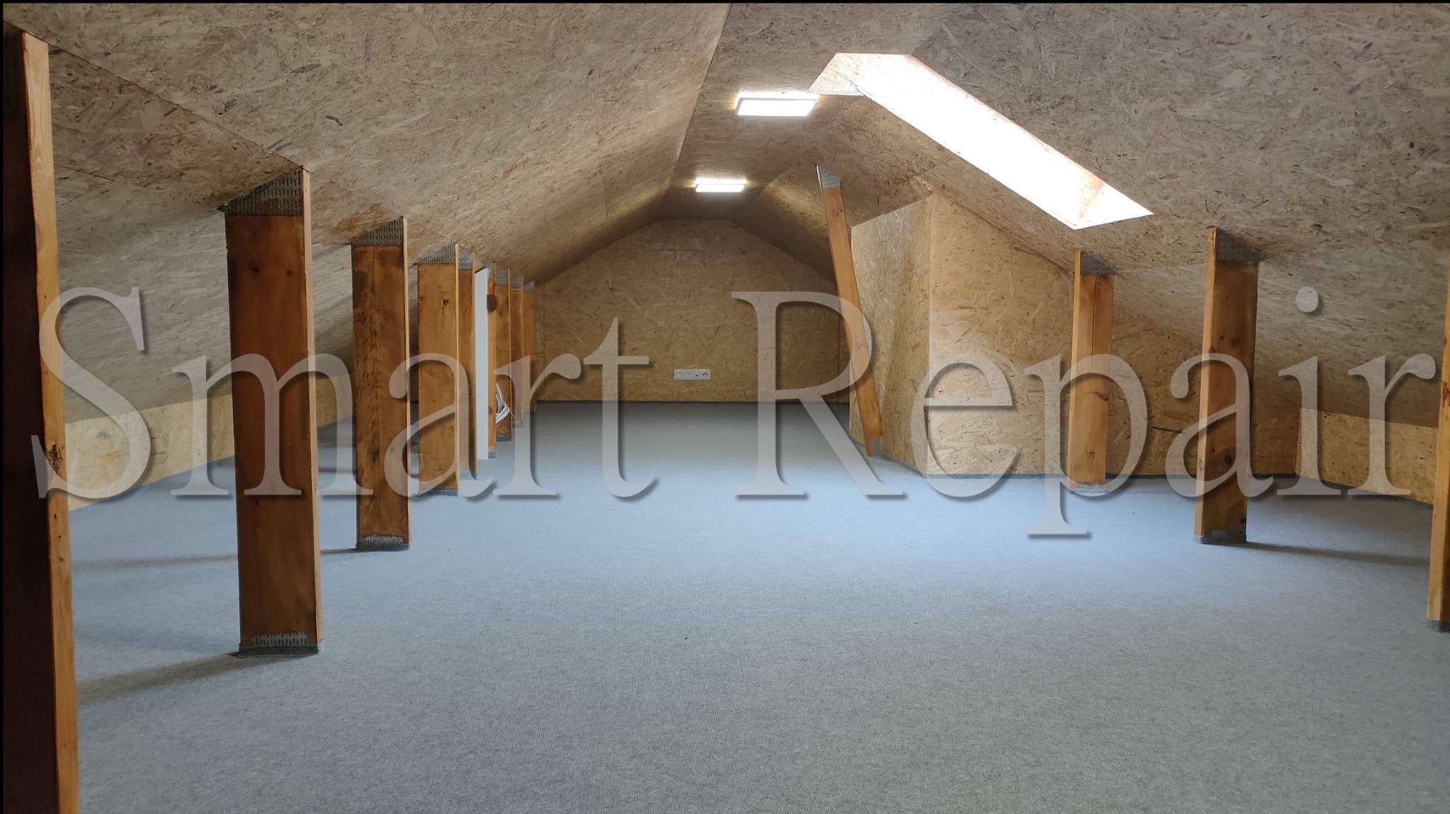 Wykończone poddasze z drewnianą konstrukcją, oknami dachowymi i szarą wykładziną. Widoczne drewniane słupy i płyty OSB na ścianach i suficie, oświetlenie punktowe.