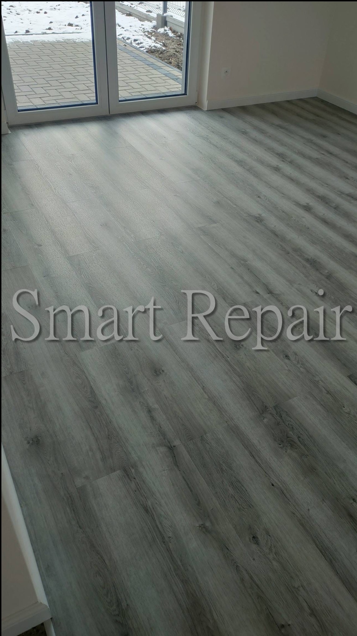 Szare panele podłogowe w nowym pokoju, widoczne z poziomu podłogi, z oknem balkonowym w tle. Na panelach widnieje napis 'Smart Repair'.