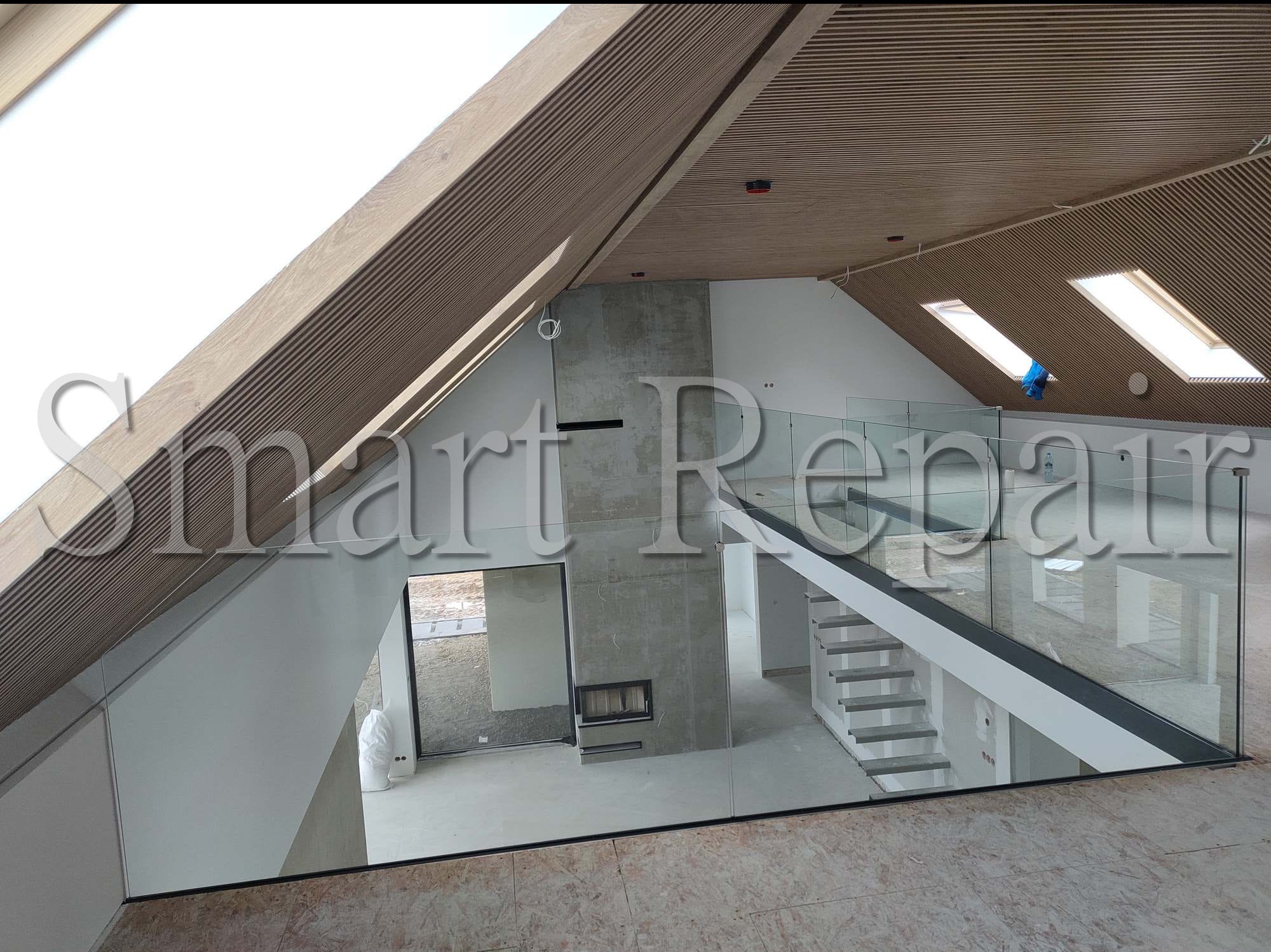 Wnętrze domu w stylu nowoczesnym z drewnianym sufitem, betonowym kominkiem i szklaną balustradą schodów. Widok z góry na przestrzeń w trakcie wykańczania.