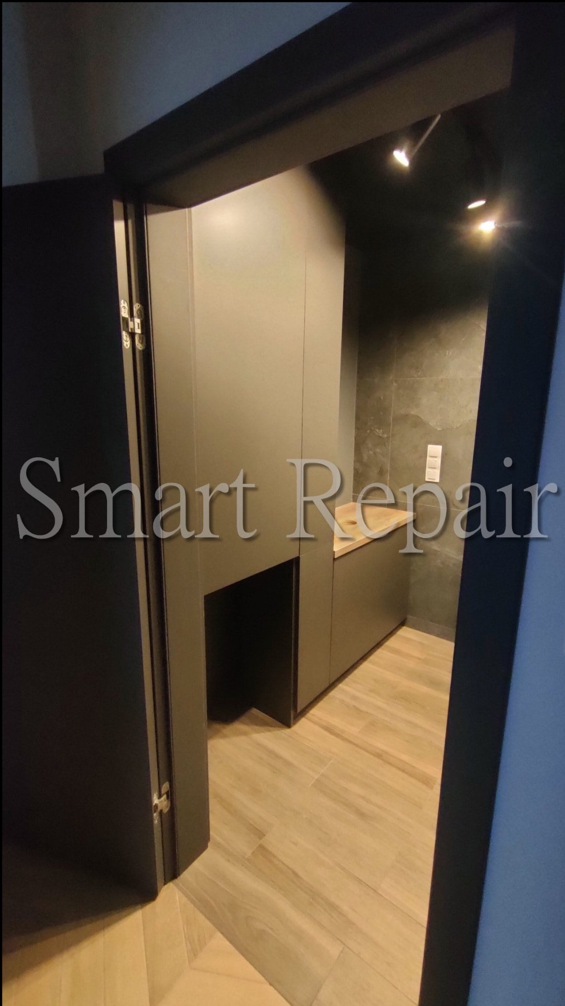 Nowoczesna garderoba w odcieniach szarości, widoczna zza otwartych drzwi. Minimalistyczny design z drewnianym blatem i subtelnym oświetleniem punktowym.