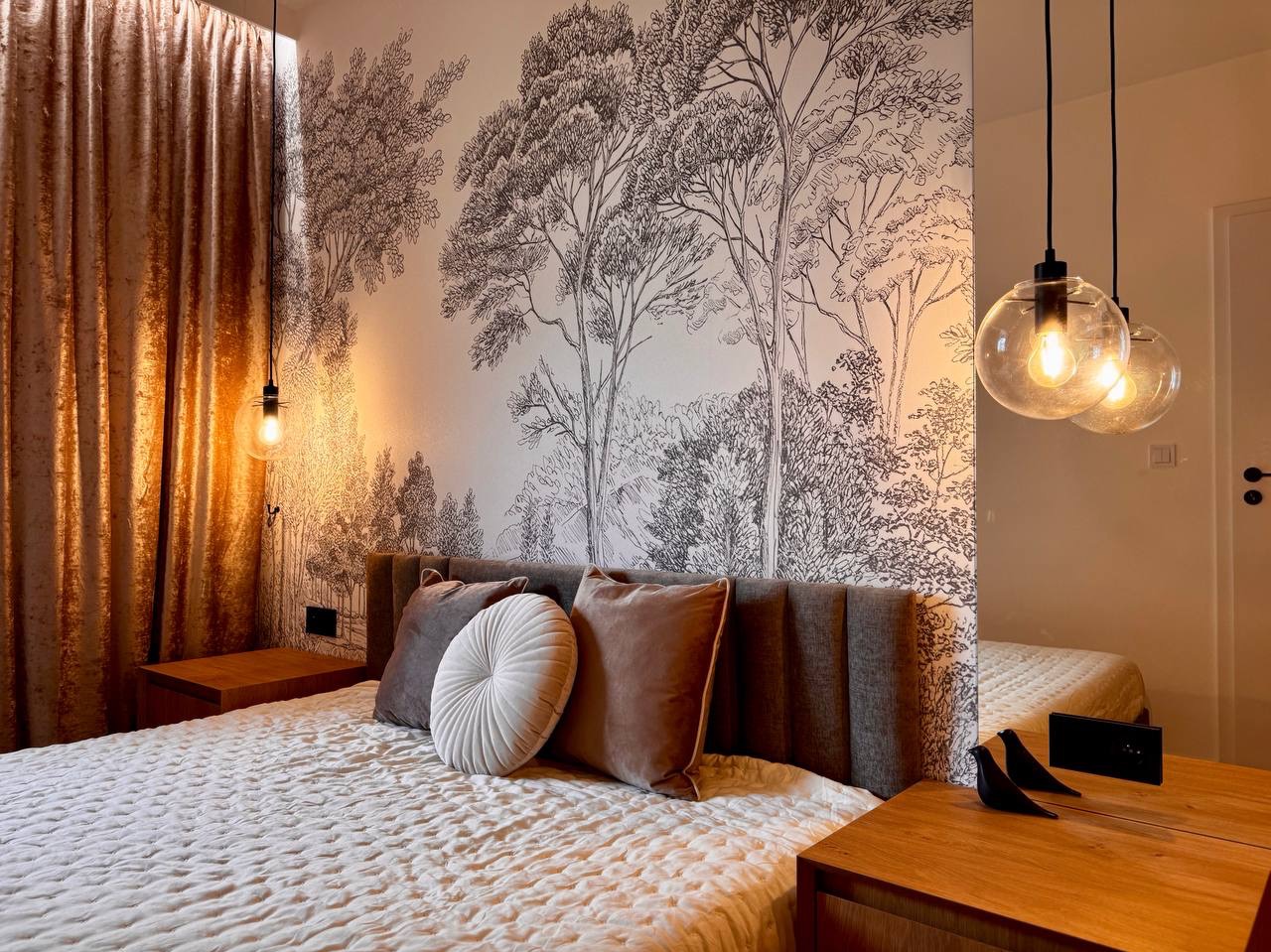 Sypialnia z tapetą w motywie leśnym, okrągłą poduszką i lampami wiszącymi. Widoczne drewniane szafki nocne i zasłony w ciepłych odcieniach brązu.