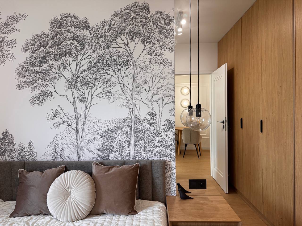 Nowoczesna sypialnia z tapetą w motywy leśne, drewnianą zabudową i wiszącymi lampami kulami. Neutralna kolorystyka, minimalistyczny design.
