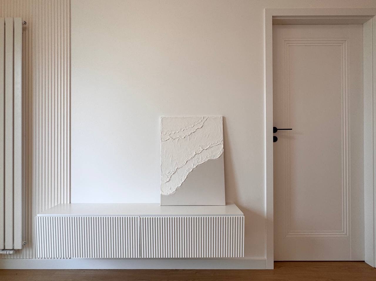 Białe wnętrze z dekoracyjną ścianą i szafką RTV z lamelami. Na szafce stoi obraz z fakturą. Widoczny fragment drzwi z czarną klamką. Minimalistyczny design.