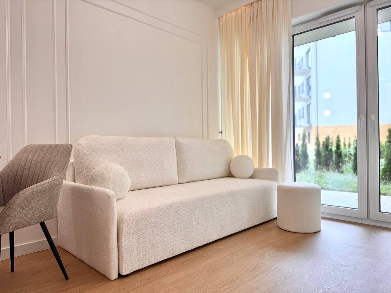 Jasny salon z sofą bouclé, pufem i krzesłem. Widok na zieleń za oknem. Minimalistyczny wystrój, drewniana podłoga, kremowe zasłony i białe ściany z dekoracyjną sztukaterią.