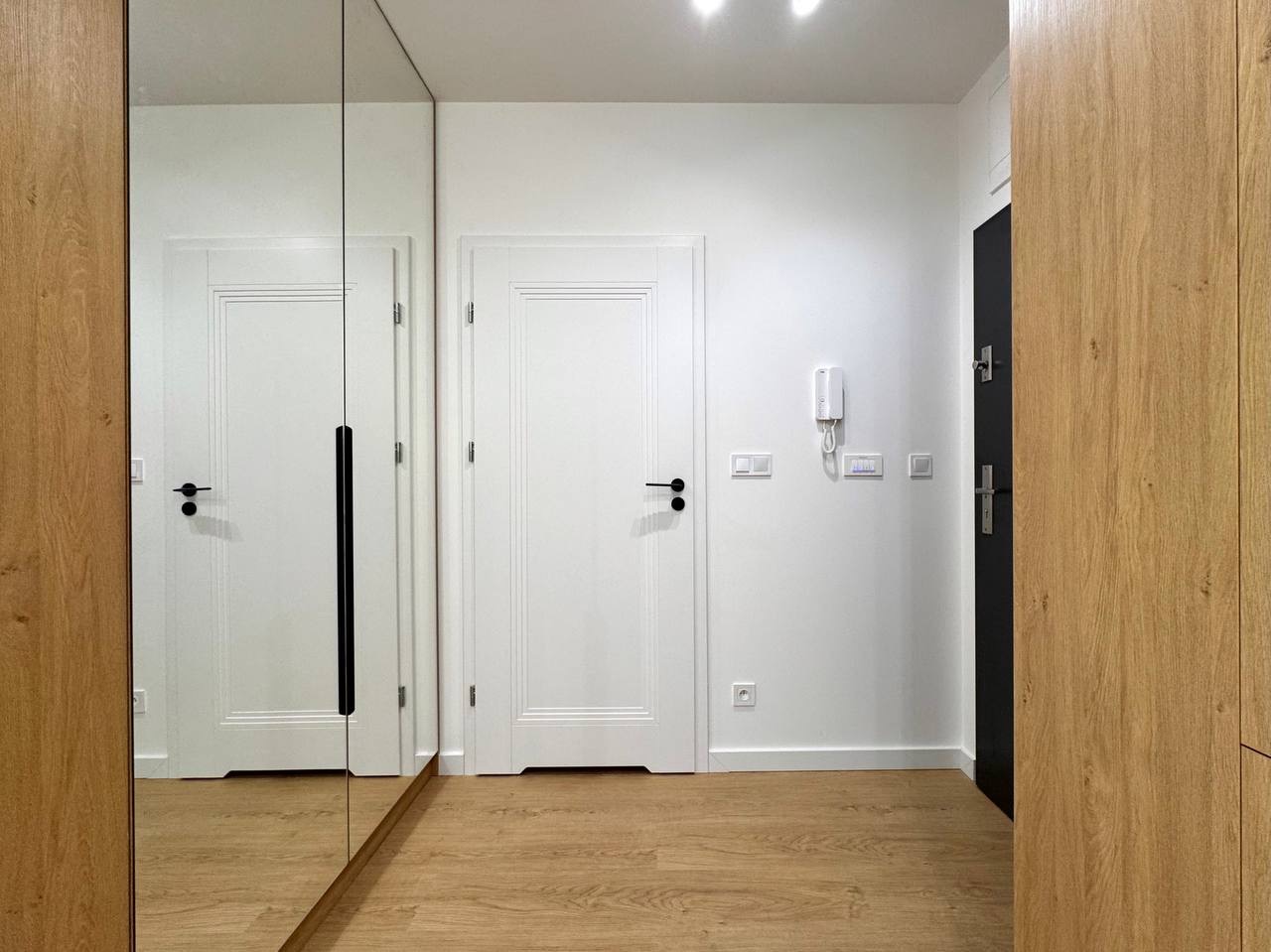 Wykończony korytarz w Jeleniej Górze: białe ściany, drewniana podłoga i szafa w zabudowie z lustrem. Minimalistyczny design, widoczny domofon i drzwi wejściowe.