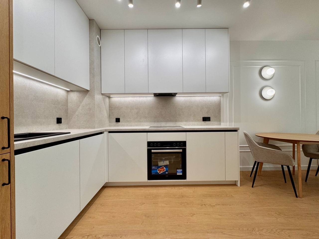 Nowoczesna, jasna kuchnia z białymi szafkami, drewnianą podłogą i okrągłym stołem. Oświetlenie LED pod szafkami dodaje elegancji. Minimalistyczny design.