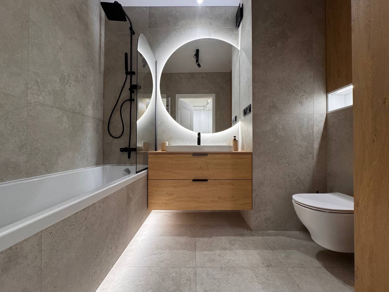 Nowoczesna łazienka z podświetlanym, okrągłym lustrem nad drewnianą szafką, szara glazura, czarna armatura i biała wanna. Widoczna toaleta i oświetlenie podłogowe.