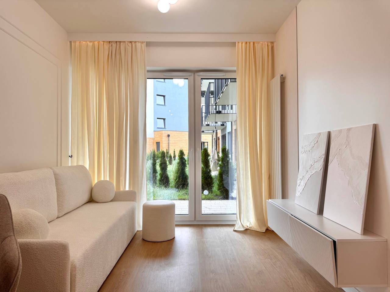 Jasne wnętrze pokoju z kremową sofą, pufem i szafką RTV. Okno z widokiem na zieleń. Minimalistyczny wystrój, drewniana podłoga i obrazy na ścianie.