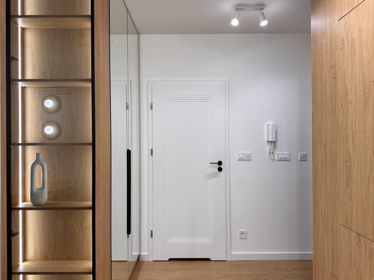 Nowoczesny przedpokój z szafą w zabudowie, oświetleniem LED i lustrem. Minimalistyczny design, białe ściany, drewniane wykończenia i dekoracyjny wazon na półce.