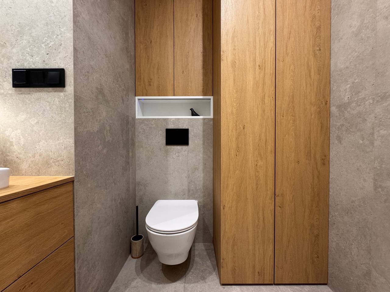 Nowoczesna łazienka z szarą płytką, zabudową z drewna i podwieszaną toaletą. Minimalistyczny design z akcentami czerni i dekorem w postaci figurki ptaka.