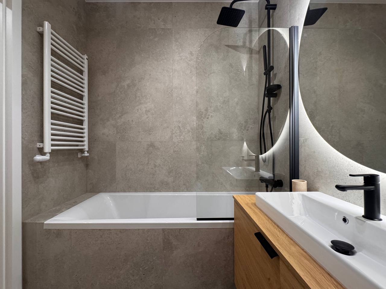 Nowoczesna łazienka z wanną, grzejnikiem drabinkowym i umywalką na drewnianej szafce. Okrągłe lustro z podświetleniem LED i czarna armatura.