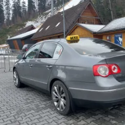 Szary samochód osobowy z żółtym napisem TAXI na dachu, zaparkowany na tle drewnianych budynków i skoczni narciarskiej w zimowej scenerii.