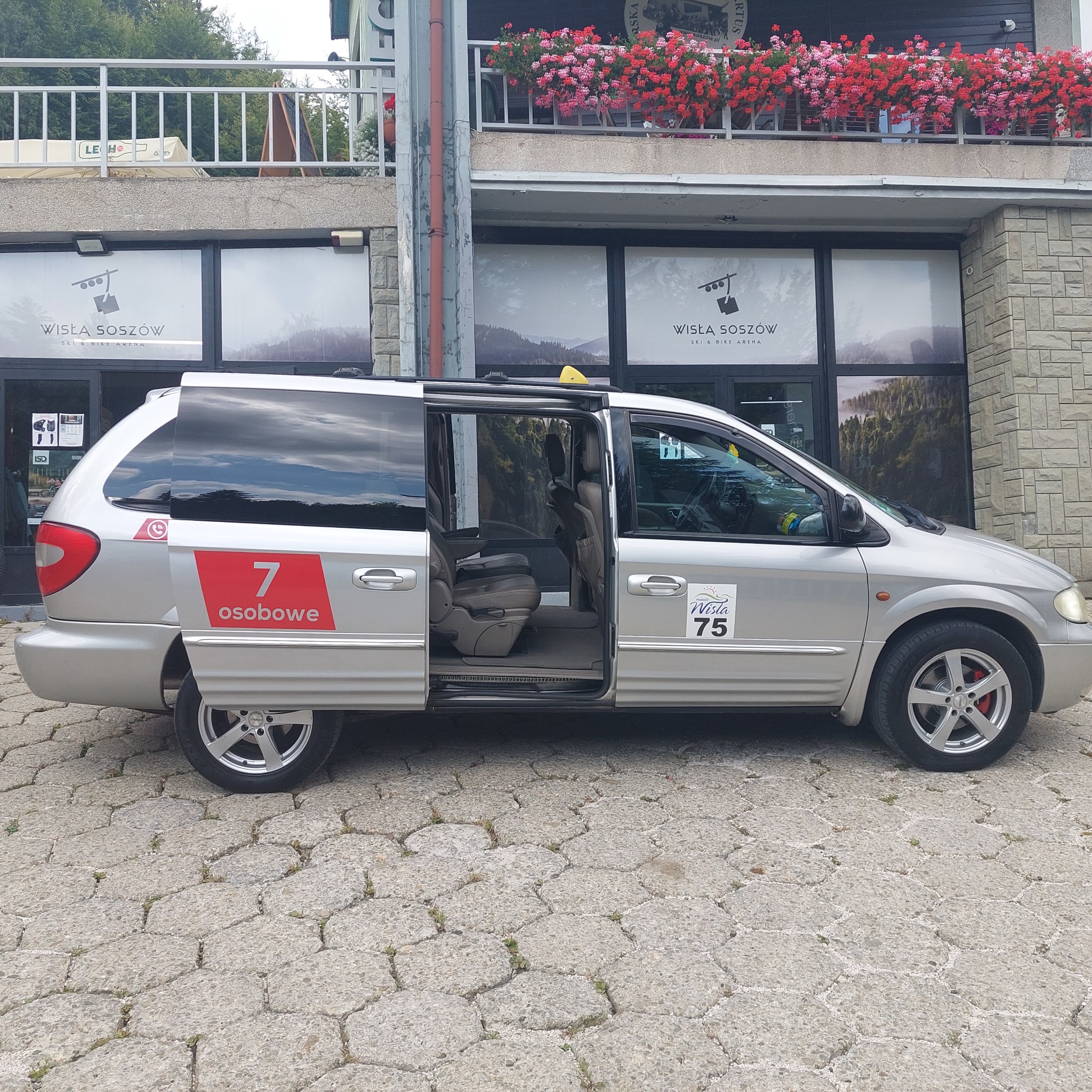 Srebrny van z otwartymi drzwiami, oznaczony numerem 75 i logo Wisła, zaparkowany przed budynkiem Wisła Soszów Ski & Bike Arena, gotowy do przewozu 7 pasażerów.