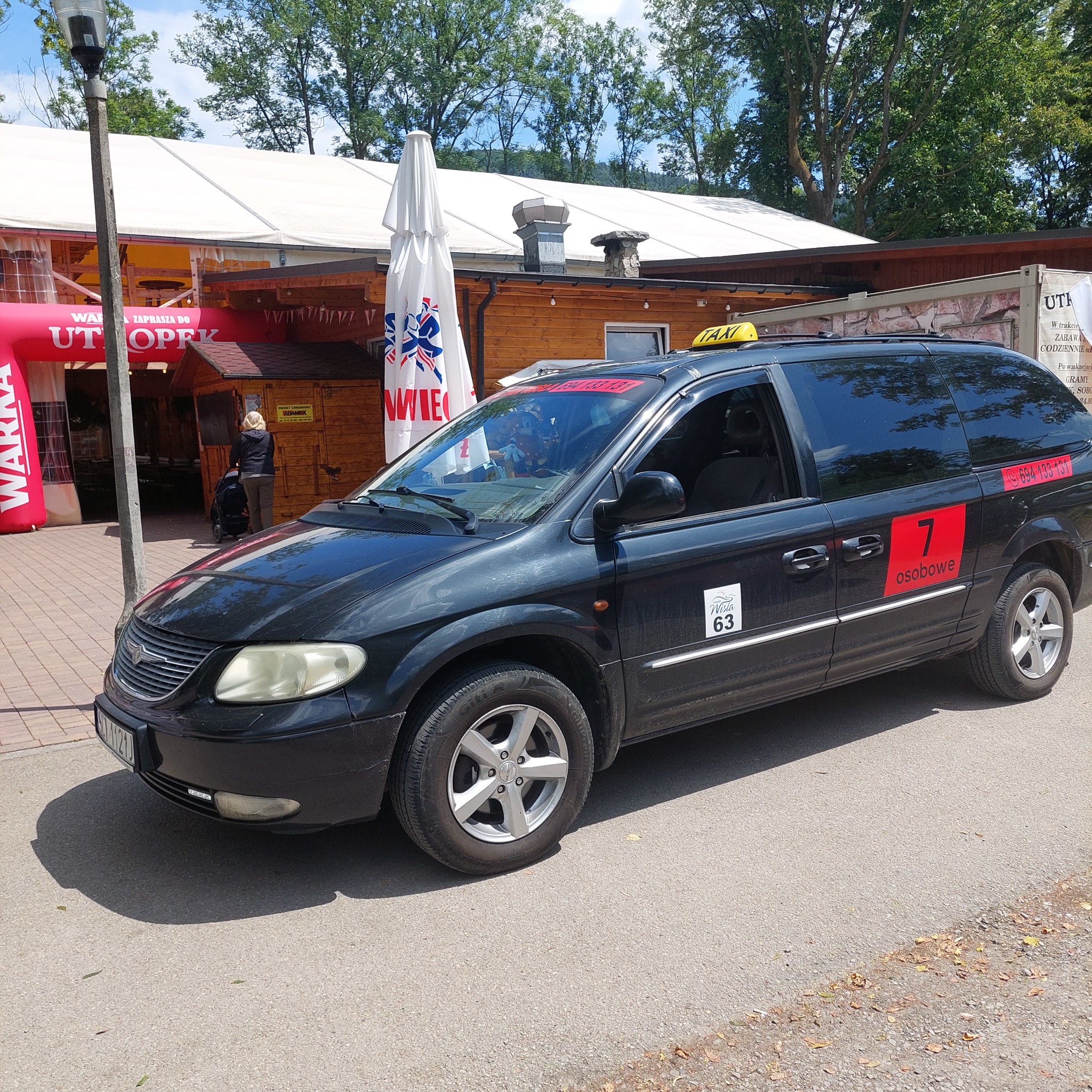 Czarny van taxi z oznaczeniami '7 osobowe' i numerem telefonu na tle drewnianej budowli z parasolem. Ujęcie z boku na ulicy.