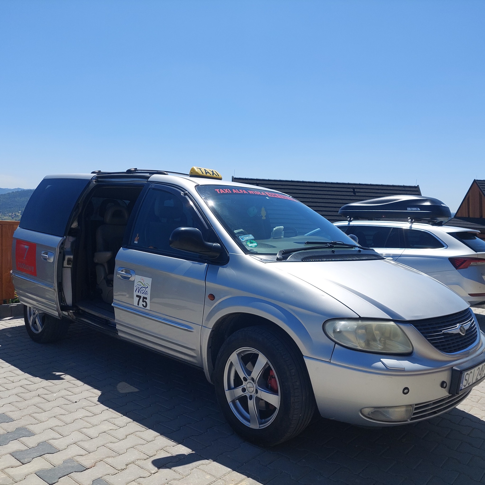 Srebrny van taxi z otwartymi drzwiami na tle błękitnego nieba i gór. Na dachu żółty szyld TAXI, na szybie napis 'TAXI ALFA WISŁA'.