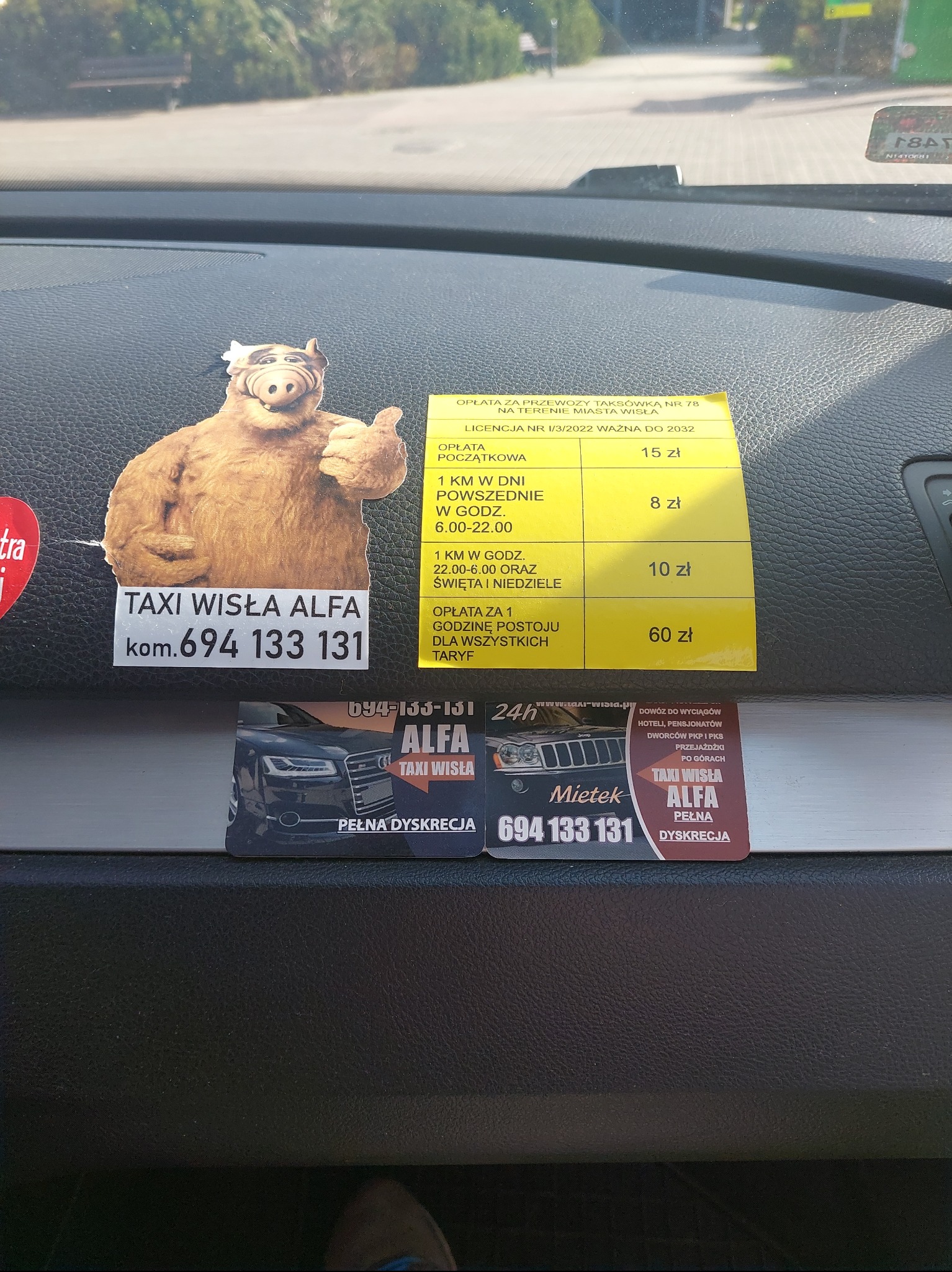 Deska rozdzielcza taksówki z naklejką Alfa Taxi Wisła, cennikiem i wizytówkami. Naklejka z ALF-em dodaje unikalnego charakteru. Numer telefonu widoczny.