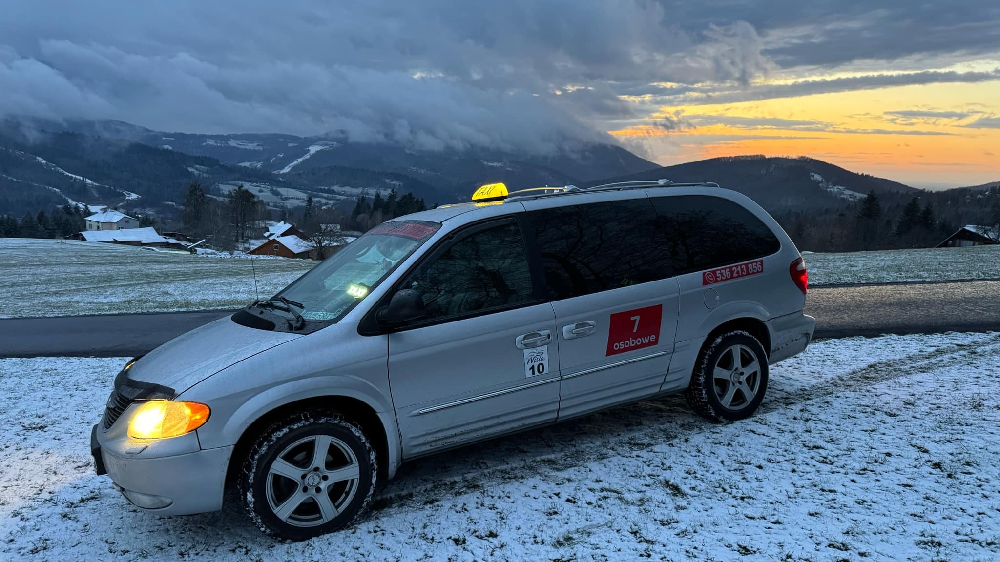 Srebrny van taxi na tle zimowego krajobrazu górskiego o zmierzchu. Widoczny numer telefonu i oznaczenie '7 osobowe'.