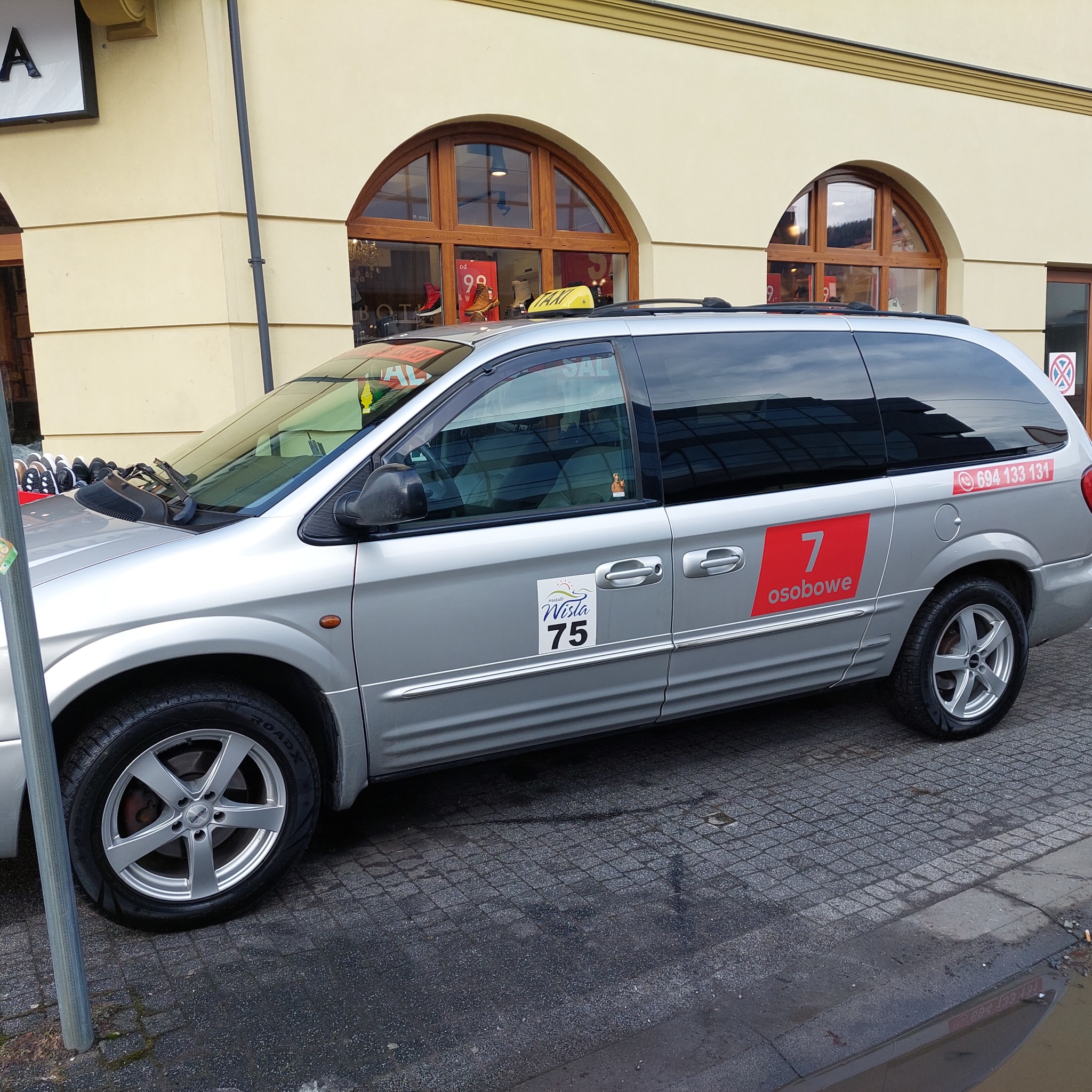Srebrny van taxi z numerem bocznym 7 i napisem 'osobowe' na tle budynku z oknami w kształcie łuków i chodnika z kostki. Widoczny znak taxi na dachu pojazdu.