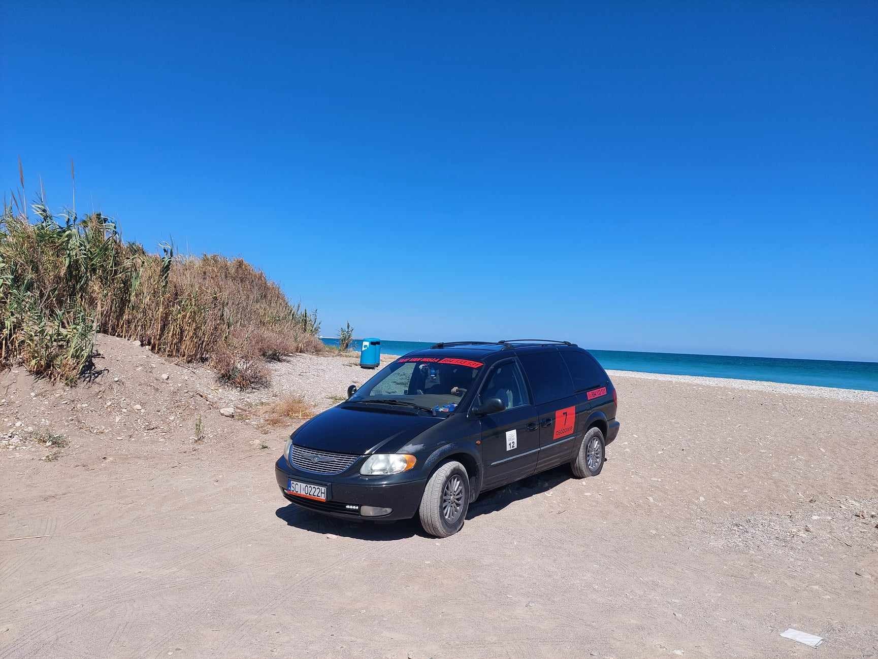 Czarny van z numerem 12 i czerwonymi napisami na dachu, zaparkowany na piaszczystej plaży z błękitnym niebem i morzem w tle. Tablica rejestracyjna widoczna.