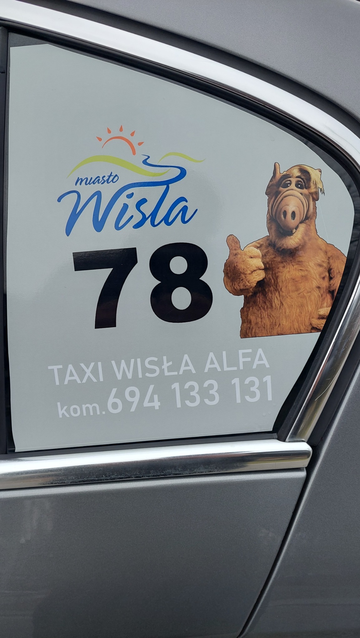 Naklejka reklamowa na szybie taksówki w Wiśle z numerem 78, logo miasta i postacią Alfa z podniesionym kciukiem. Kontakt: 694 133 131.