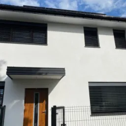 Biały dom z czarnymi żaluzjami okiennymi i drzwiami z drewna, widok z zewnątrz, ogrodzenie panelowe w tle, błękitne niebo z chmurami.