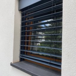 KrisLine Krzysztof Demianiuk - Szare listwy żaluzji fasadowej zamontowane w oknie budynku z białą, chropowatą elewacją. Widoczne odbicie drzew w szybie.