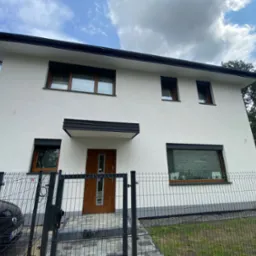 Biały dom jednorodzinny z brązowymi oknami i czarnymi roletami zewnętrznymi, widziany zza ogrodzenia. Roleta na parterze jest opuszczona.