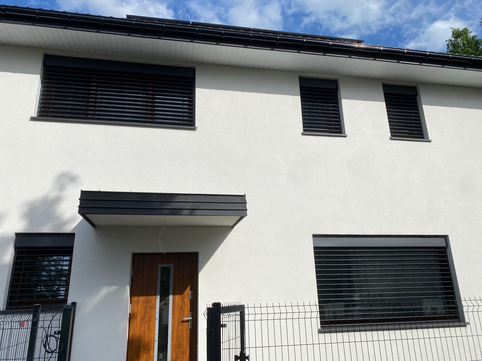 Biały dom z czarnymi żaluzjami okiennymi i drzwiami z drewna, widok z zewnątrz, ogrodzenie panelowe w tle, błękitne niebo z chmurami.