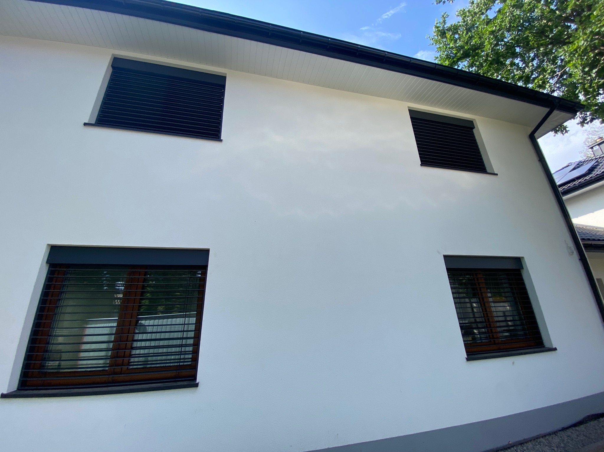 Biały dom z czterema oknami, z których dwa mają opuszczone, czarne żaluzje fasadowe, a dwa są otwarte, ukazując drewniane ramy okienne.