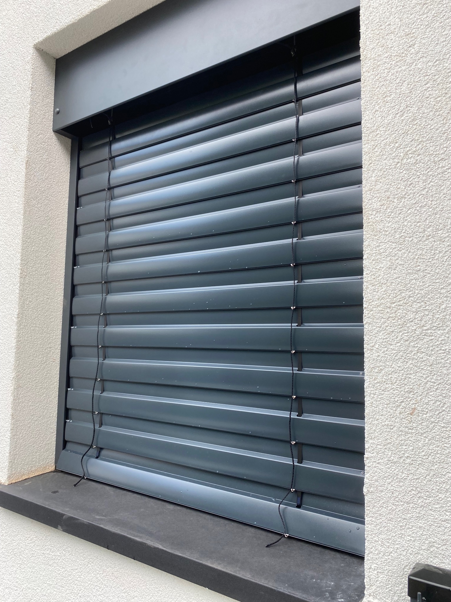 Zewnętrzna żaluzja fasadowa w kolorze grafitowym, zamontowana na oknie budynku z białą, chropowatą elewacją. Widoczny fragment parapetu i linki sterujące.