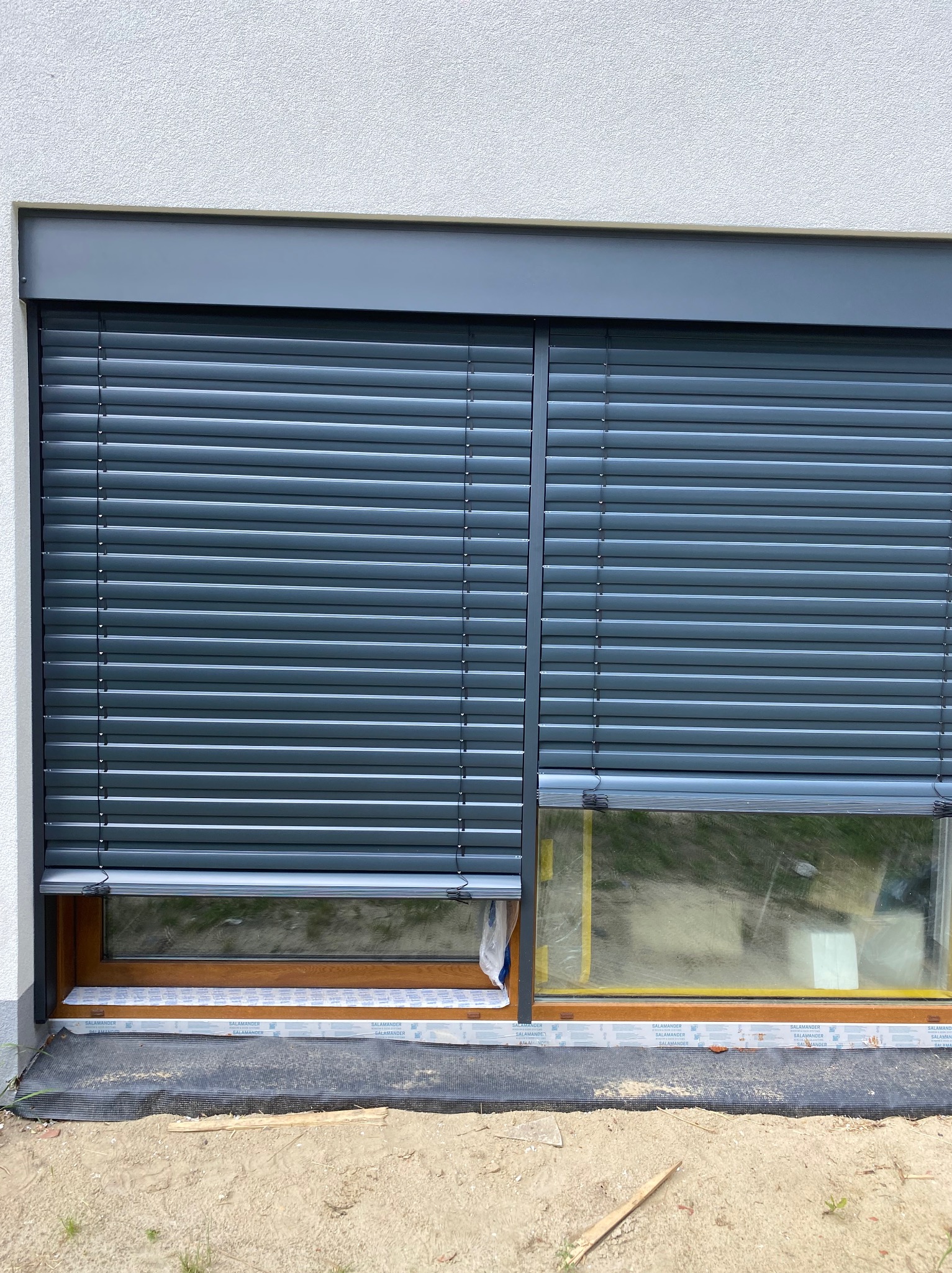 Szare, częściowo opuszczone żaluzje fasadowe na oknie budynku z widocznym fragmentem ramy okiennej. Ujęcie z bliska, w tle biała elewacja budynku.