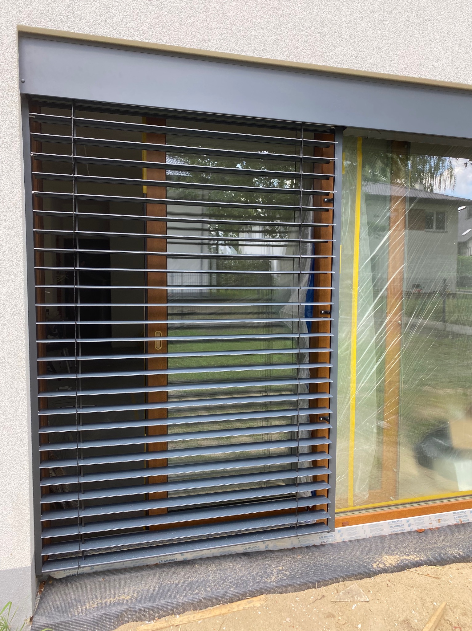 Zamknięte szare żaluzje fasadowe z poziomymi lamelami na oknie budynku w trakcie wykończenia. Widoczna folia ochronna na sąsiednim oknie.