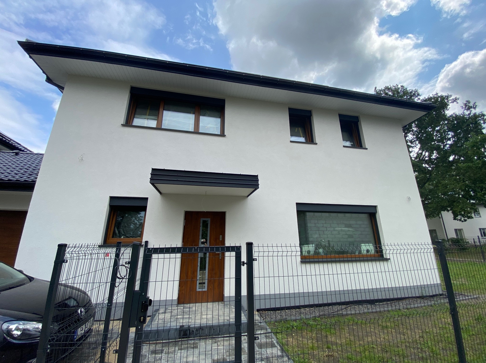 Biały dom jednorodzinny z brązowymi oknami i czarnymi roletami zewnętrznymi, widziany zza ogrodzenia. Roleta na parterze jest opuszczona.