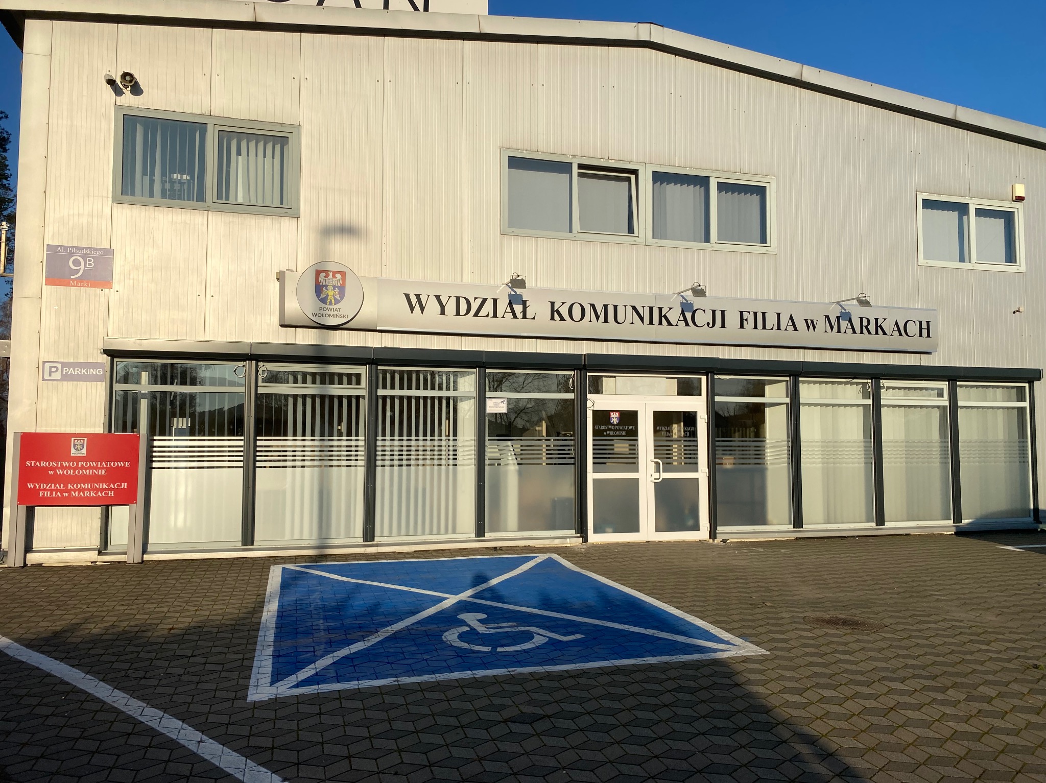 Budynek Wydziału Komunikacji w Markach z widocznymi oknami zabezpieczonymi poziomymi żaluzjami wewnętrznymi. Na zewnątrz oznakowane miejsce parkingowe dla niepełnosprawnych.
