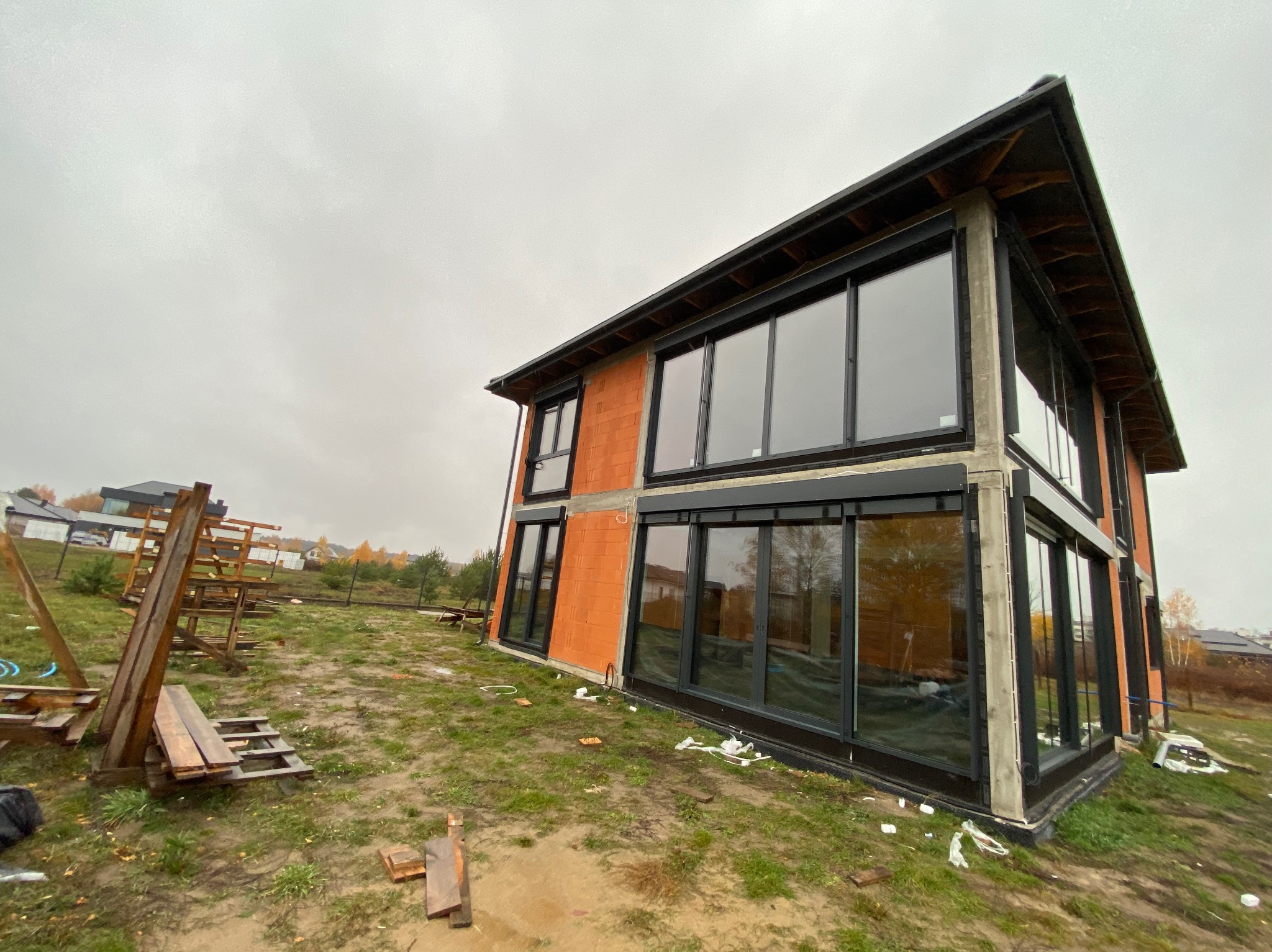 Nowoczesny dom w trakcie budowy z zamontowanymi oknami i czarnymi ramami. Widoczne rusztowania i materiały budowlane na zewnątrz. Pochmurne niebo w tle.