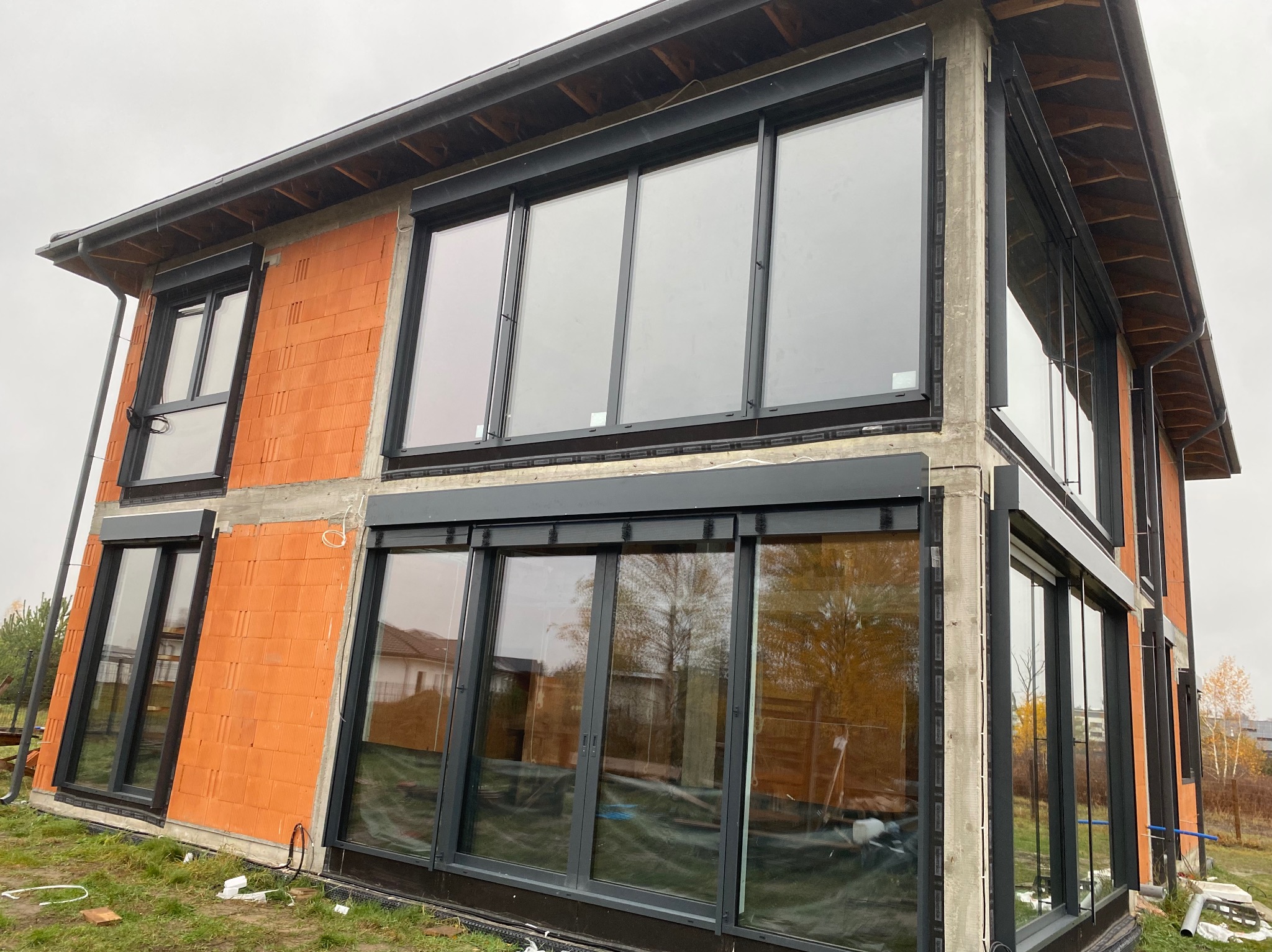 Nowoczesny dom w trakcie budowy z zamontowanymi, grafitowymi roletami zewnętrznymi na oknach. Widok z zewnątrz, w tle jesienne drzewa.