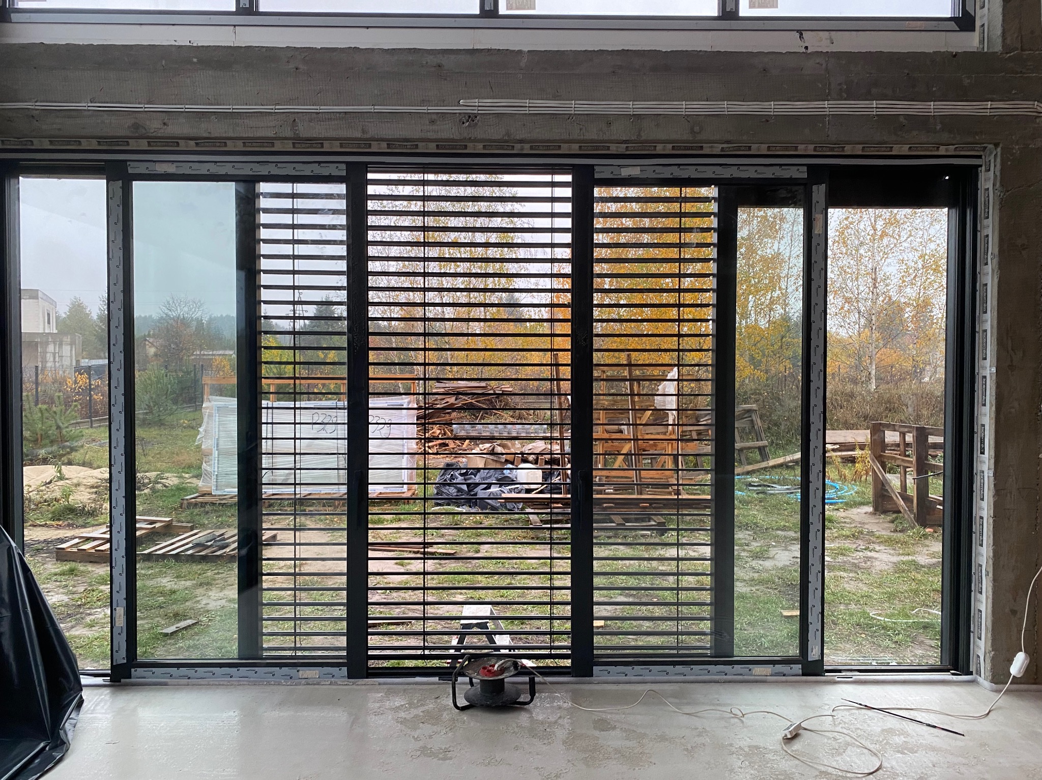 Zamontowane żaluzje w oknie przesuwnym z widokiem na jesienny ogród. Widoczne narzędzia montażowe na podłodze. Surowy stan wykończenia wnętrza.