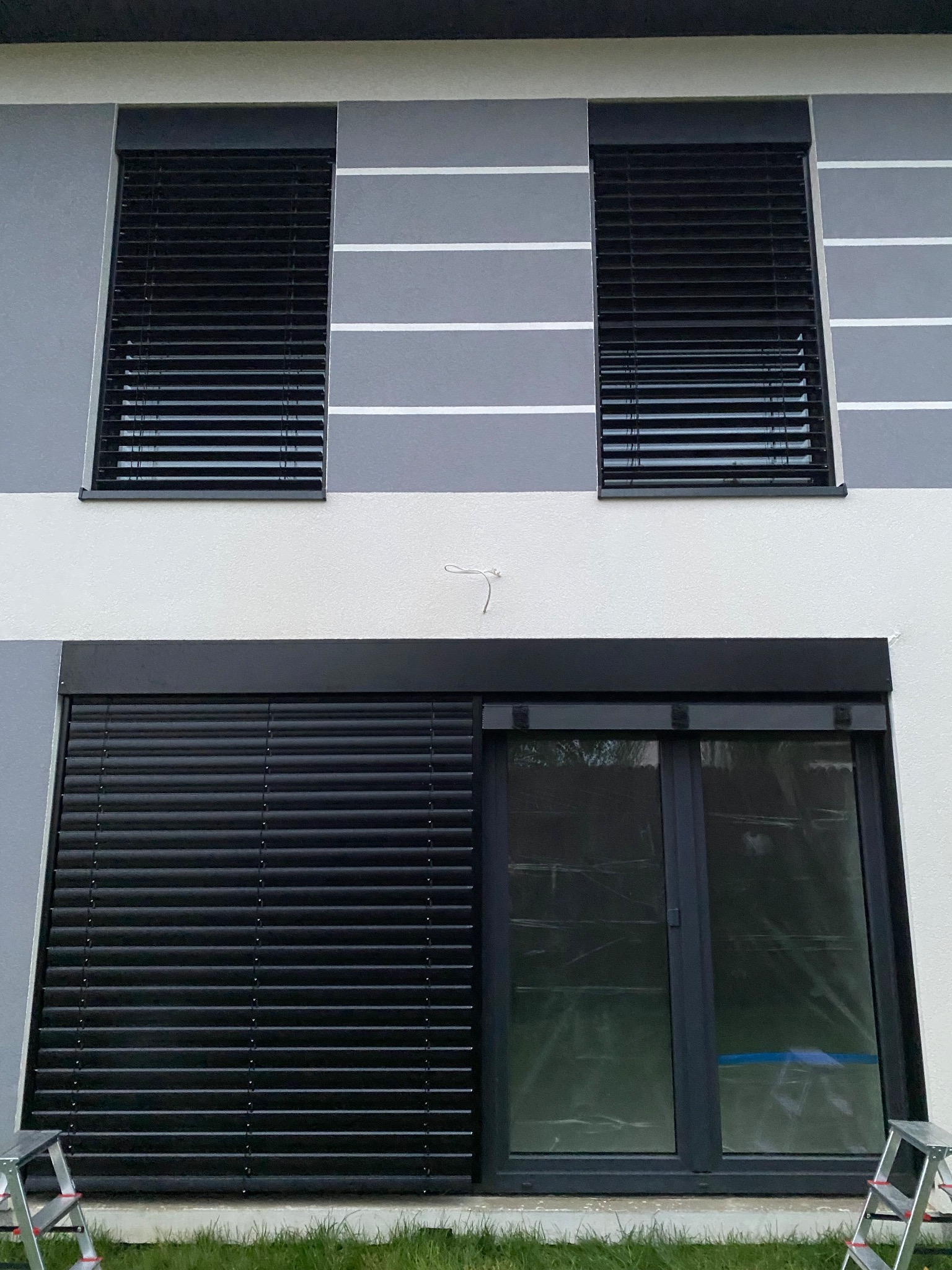 Elewacja domu z dwoma oknami z czarnymi żaluzjami i dużym oknem przesuwnym z czarną roletą. Widoczne drabiny sugerują niedawny montaż.