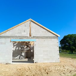 Betonowa Moc - Wznoszenie ścian parterowego domu z betonu komórkowego. Widoczne drewniane elementy więźby dachowej i rusztowanie w otworze drzwiowym. Słoneczny dzień.