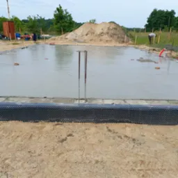 Zalany fundament z wystającymi prętami zbrojeniowymi, zabezpieczony folią kubełkową. Prace budowlane w Szprotawie. W tle hałda piachu i zieleń.