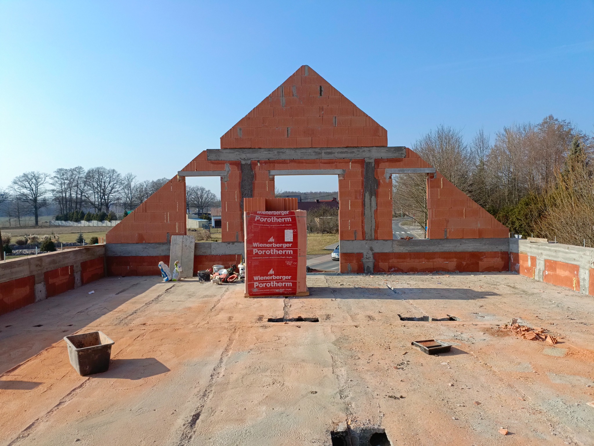 Budowa domu z czerwonej cegły Porotherm. Widoczne fragmenty ścian z otworami na okna i drzwi oraz paleta cegieł. W tle drzewa i błękitne niebo.