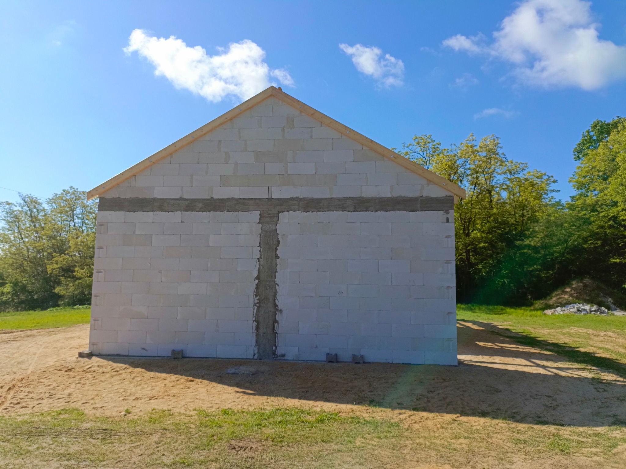 Wzniesiony do stanu surowego otwartego budynek z białych bloczków, widoczne zbrojenia w ścianach, drewniana konstrukcja dachu, błękitne niebo w tle.