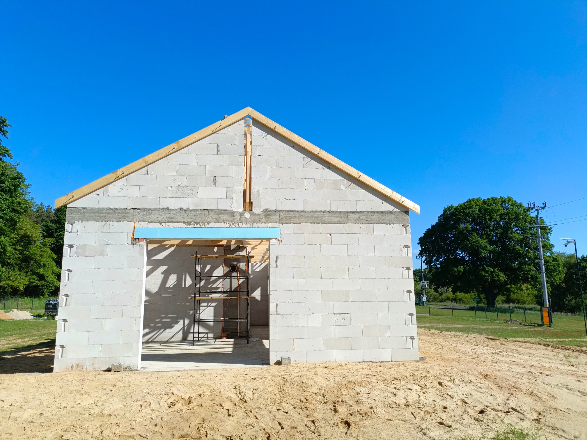 Wznoszenie ścian parterowego domu z betonu komórkowego. Widoczne drewniane elementy więźby dachowej i rusztowanie w otworze drzwiowym. Słoneczny dzień.