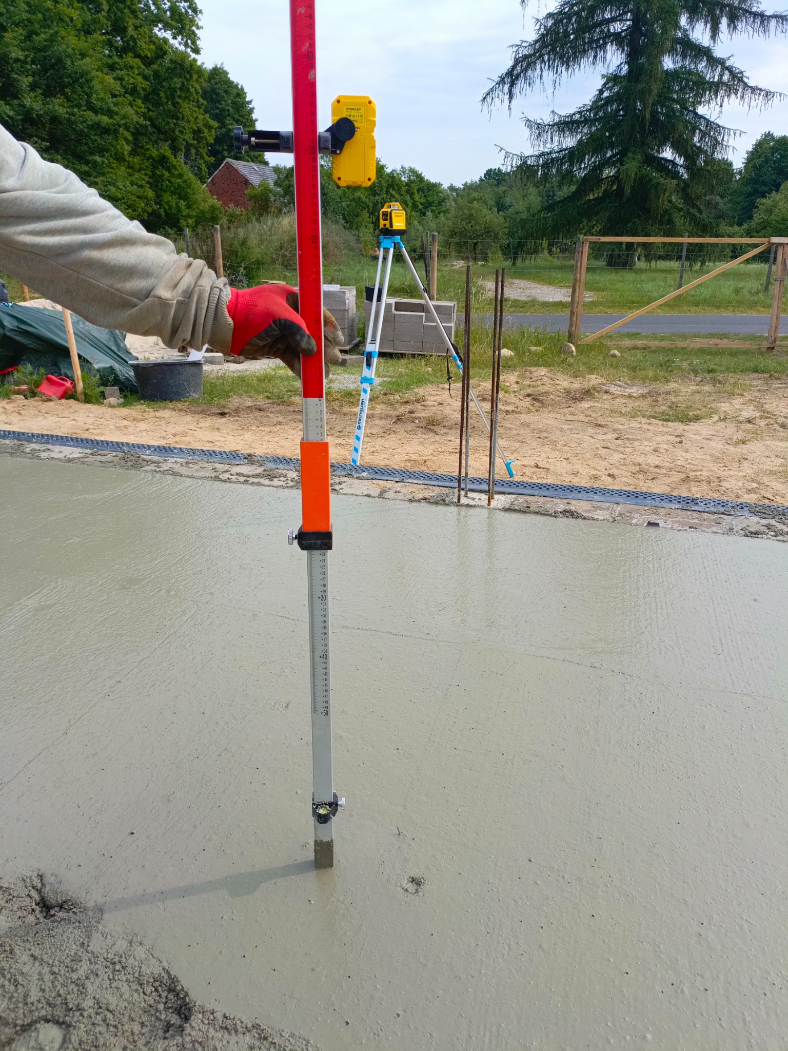 Wyrównywanie wylewki betonowej z użyciem łaty niwelacyjnej i niwelatora laserowego. Widoczne zbrojenie i odpływ liniowy. Prace budowlane na zewnątrz.