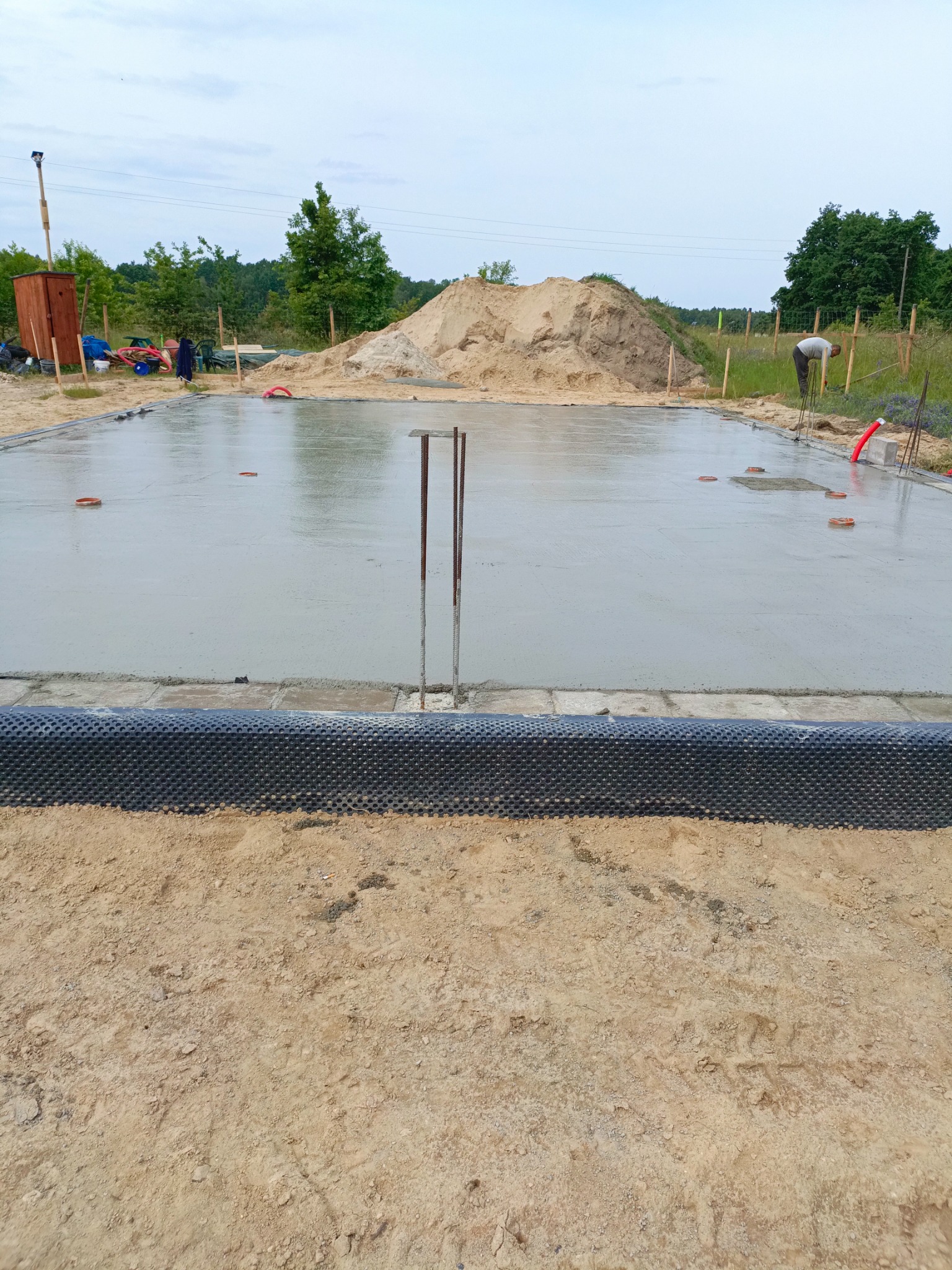 Zalany fundament z wystającymi prętami zbrojeniowymi, zabezpieczony folią kubełkową. Prace budowlane w Szprotawie. W tle hałda piachu i zieleń.