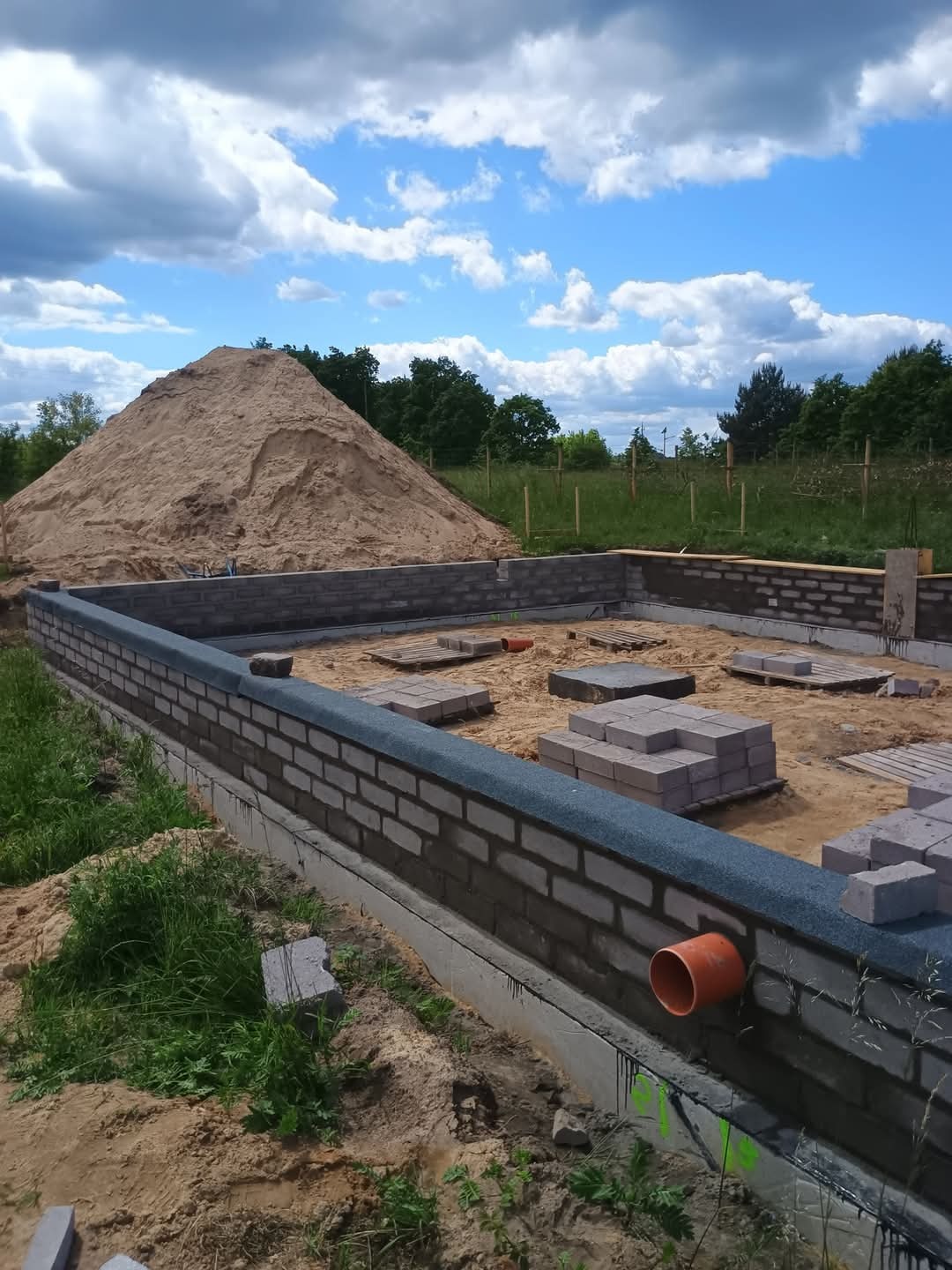 Fundamenty z bloczków betonowych na piasku, widoczna izolacja przeciwwilgociowa i stos piasku w tle. Prace murarskie w toku, widok na częściowo ukończone ściany fundamentowe.