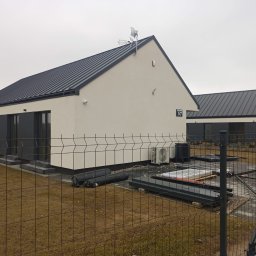 FSDOMY - Nowoczesny, parterowy dom z ciemnym dachem i jasną elewacją, widoczny zza ogrodzenia. Na posesji elementy instalacyjne i klimatyzator. Tabliczka z numerem 57.