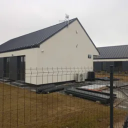 Nowoczesny, parterowy dom z ciemnym dachem i jasną elewacją, widoczny zza ogrodzenia. Na posesji elementy instalacyjne i klimatyzator. Tabliczka z numerem 57.