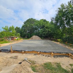 FSDOMY - Wylana płyta fundamentowa z drewnianym szalunkiem na piaszczystym terenie, widoczny biały samochód ciężarowy i sterta ziemi w tle. Słoneczny dzień.