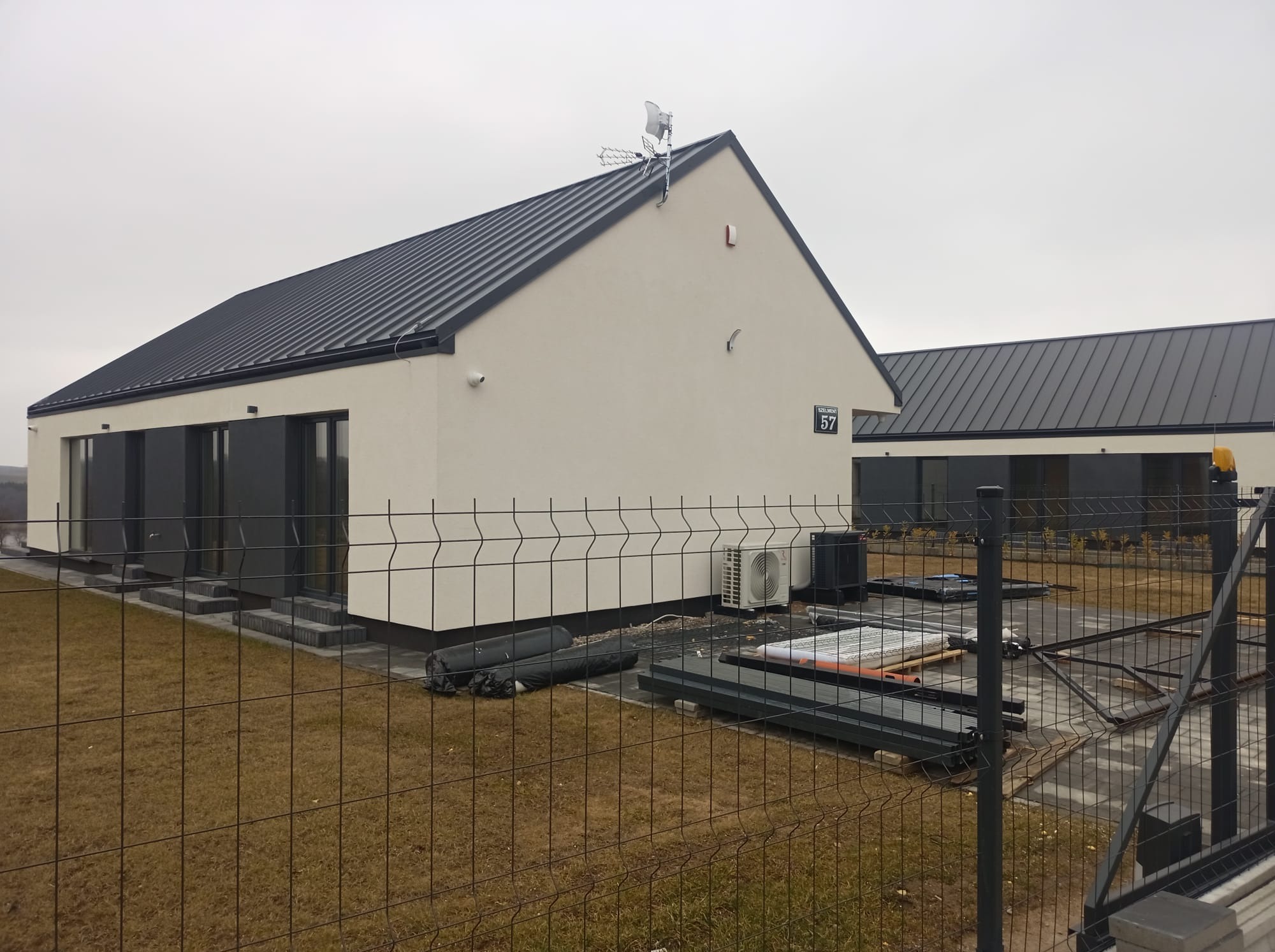 Nowoczesny, parterowy dom z ciemnym dachem i jasną elewacją, widoczny zza ogrodzenia. Na posesji elementy instalacyjne i klimatyzator. Tabliczka z numerem 57.